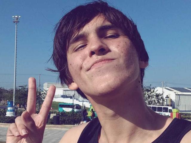 AngelW with messy dark hair, fair skin, and small zits on face, making a "peace" sign, wearing a black tank top, smiling slightly under clear blue sky.