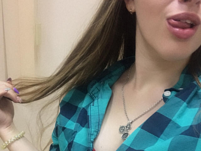 Dooris with pouty pink lips, blue-green checkered shirt, necklace, holding brown hair, purple nails, gold bracelet, and small earrings, showing cleavage.