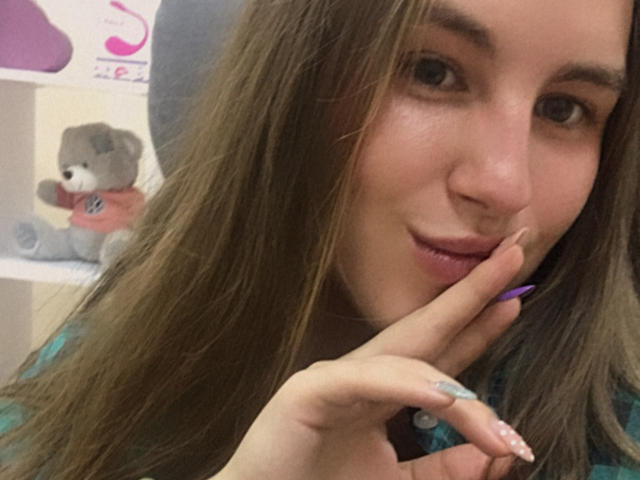 Dooris with long brown hair, subtle smile, and purple nails, touching her slightly parted lips with her finger. A cute gray stuffed animal with a pink shirt is in the background.