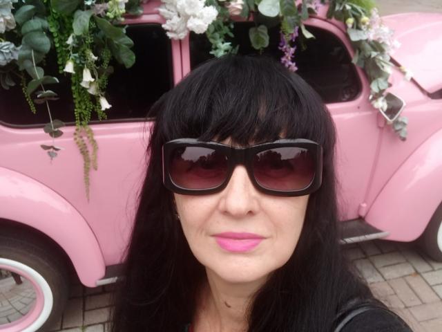 MelanyBanks with black hair, pink lipstick, and dark sunglasses, standing in front of a pink vintage car decorated with green and white flowers. Boobs not visible.