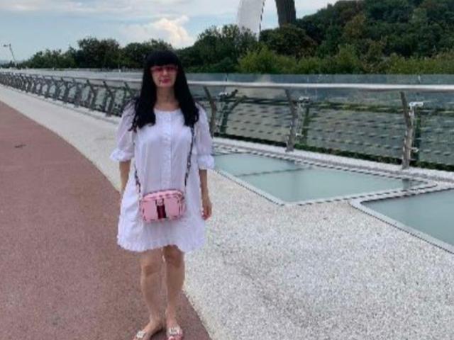 MelanyBanks has black hair, wears a white sheer dress, pink crossbody bag, and pink glasses, standing on a gravel path with a metal railing beside a waterway.