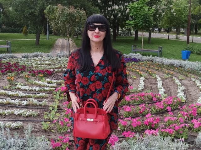 Melany Banks, avec ses longs cheveux noirs, portant des lunettes de soleil noires, une robe fleurie rouge et un sac à main rouge, se tient devant des parterres de fleurs colorés. Elle porte du rouge à lèvres et a une belle silhouette..