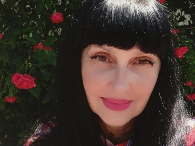 MelanyBanks with black hair, bangs, and pink lipstick, smiling at the camera, has brown eyes, and is surrounded by red roses in the background. Her tits are not visible.