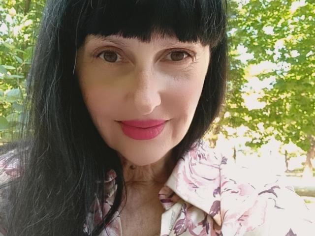 Melany Banks tiene el pelo negro, ojos marrones y lápiz labial rosa. Lleva una camisa con estampado floral, y sus senos son parcialmente visibles. Está sonriendo, y se ve linda..
