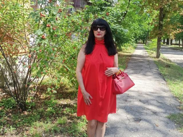 MelanyBanks with black hair, wearing a red sleeveless dress, red bag, and sunglasses, standing on a sidewalk, hand on dress, green leafy background, sunny day.