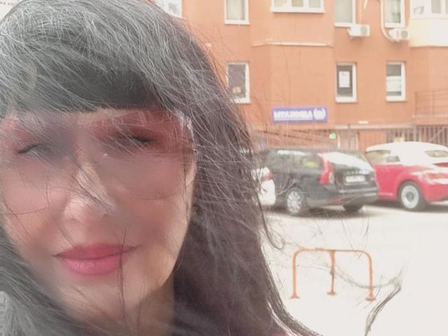 Melany Banks, con el cabello negro despeinado, lápiz labial rojo y el rostro ligeramente borroso, está de pie frente a coches estacionados, vistiendo una camisa oscura. Sus senos no son visibles..