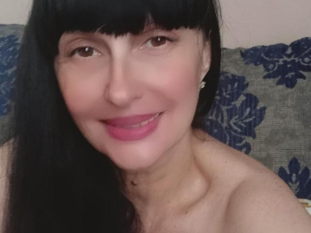 MelanyBanks has black hair, brown eyes, pink lipstick, and is smiling with her bare shoulders visible. She's wearing small earrings. Her skin is fair.