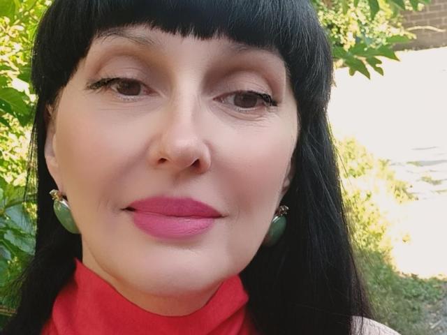 MelanyBanks has black hair, pink lipstick, green earrings, and a red turtleneck, looking at the camera with a slight smile. Her nose is small and her skin is fair.
