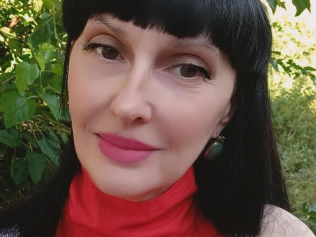 Melany Banks heeft zwart haar, roze lippenstift, groene oorbellen, een rode coltrui en een lichte glimlach, waarbij haar neus en ogen zichtbaar zijn. Haar borsten zijn niet zichtbaar..