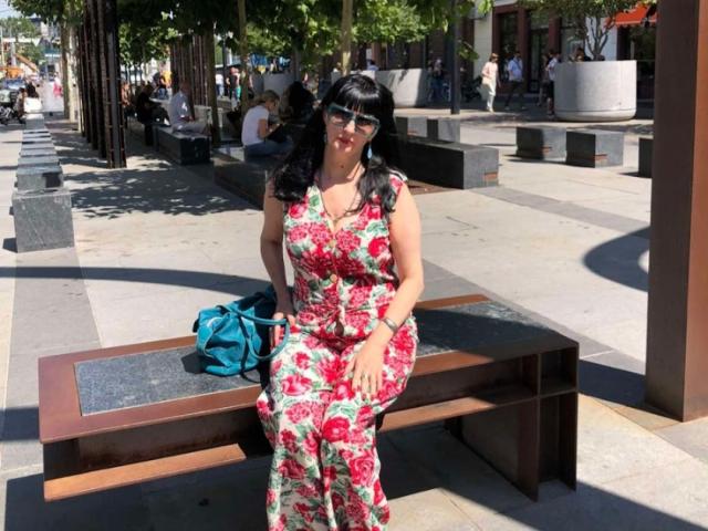Melany Banks, con cabello negro y flequillo, vestida con un vestido floral, sentada en un banco, con un bolso azul a su lado, sonriendo, iluminada por el sol, en un entorno urbano, y usando gafas de sol..