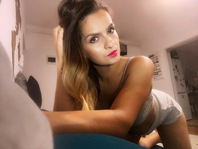 ReveMignoneX with blonde ombre hair, red lipstick, and gray underwear, leaning forward, showing her tits, with a seductive look, in a dimly lit room, hand in hair.
