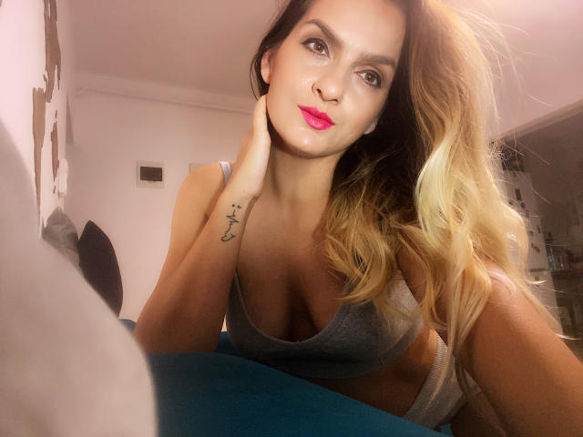 ReveMignoneX with blonde hair, gray bra, pink lipstick, and tattoo on right hand, touching cheek, looking at camera, showing small tits, and a cute face.