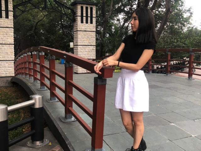 MileyWatson with black top, white skirt, black boots, holding red railing, black hair, standing, looking right, small black bracelet, stone pillars, metal railings, outdoor setting.
