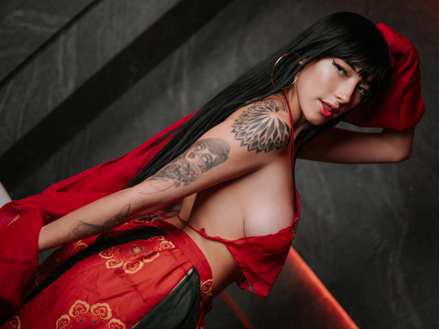 Violeta Parr, com cabelos pretos longos, vestido vermelho e brincos grandes, exibindo tatuagens e seus seios, com o braço sobre a cabeça, olhando para a câmera com batom vermelho..