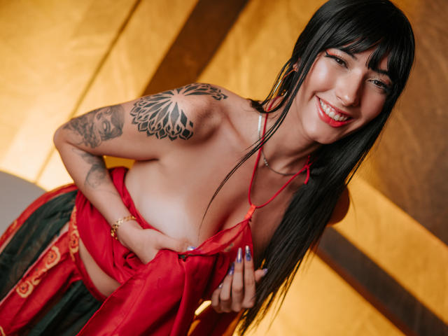 Violeta Parr mit langen, schwarzen Haaren, einem roten Halstopp, der heruntergezogen ist, um ihre Brüste zu zeigen, lächelnd, mit einem Tattoo an der rechten Schulter, einer goldenen Halskette, einem roten und grünen Rock, im Innenbereich, blickt in die Kamera..