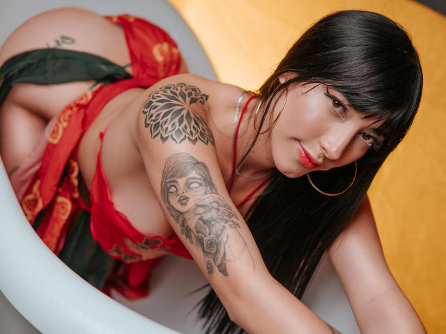 Violeta Parr, com cabelo preto, roupa íntima vermelha, tatuagens no braço e ombro, brincos grandes em formato de argola, inclinada em uma banheira branca, com um tecido vermelho e verde, olhando para a câmera com batom vermelho..