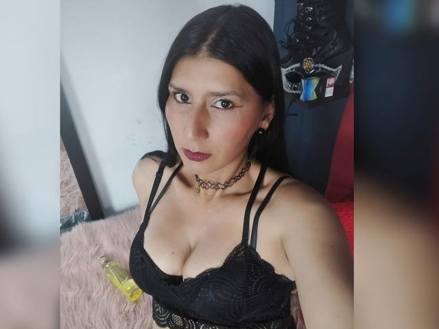 AgathaDo with long black hair, dark red lipstick, black lace bra showing her tits, choker necklace, and a yellow bottle on pink fur. She's looking at the camera.