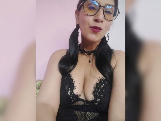 AgathaDo with black lace lingerie, showing big tits in a deep cleavage, wearing glasses, choker, and pigtails, with pink lipstick and dark makeup.
