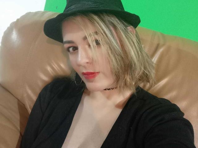 AmandaHotL with blonde hair, black hat, red lipstick, black choker, and black top, showing her tits in a brown leather chair, green background.