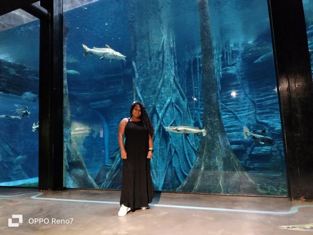 YiyaHot69 stands in front of a large aquarium, wearing a black sleeveless dress and white sneakers, with long curly hair, surrounded by fish and underwater trees.