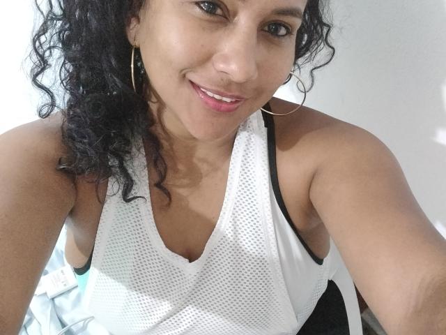 YiyaHot69 with curly black hair, wearing a white mesh sports top, showing her small tits, smiling, gold hoop earrings, light brown skin, pink lips, black strap, and upper body.