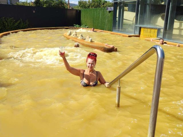 MilayaDFlow, avec ses cheveux roux, portant un bikini noir et gris, tient une boisson dans le bassin de boue jaune, agrippant l'échelle en métal, montrant ses seins, et souriant..