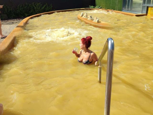 MilayaDFlow, avec des cheveux roux, tenant une boisson dans sa main droite, dans un jacuzzi jaune et turquoise, utilisant l'échelle en métal, portant un bikini noir, montrant ses seins..