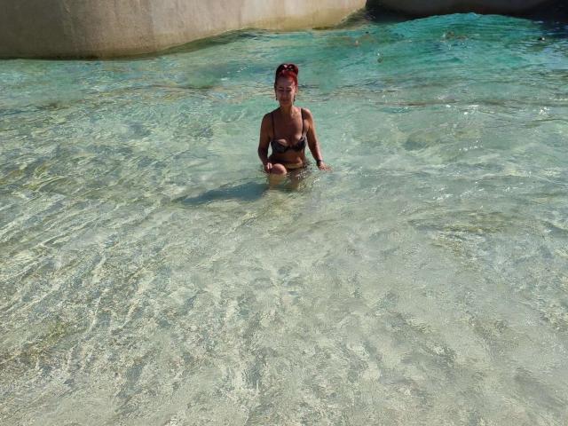 MilayaDFlow, avec de petites seins, portant un bikini noir, se tient dans une eau claire et peu profonde, ses cheveux roux attachés, souriant à l'appareil photo, dans un cadre tropical ensoleillé..