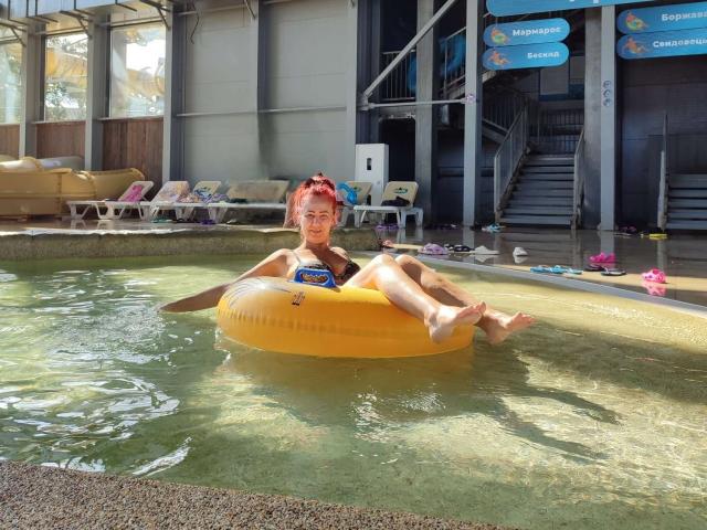 MilayaDFlow, with red hair, is in a yellow inflatable ring, floating in a pool, wearing a blue swimsuit, legs up, and a relaxed smile. Sunlight shines on her.