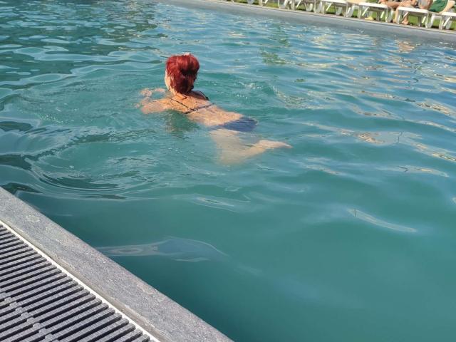 MilayaDFlow with red hair, back to camera, in blue-tinted pool, showing bare ass above water, pool edge with metal grate in foreground.