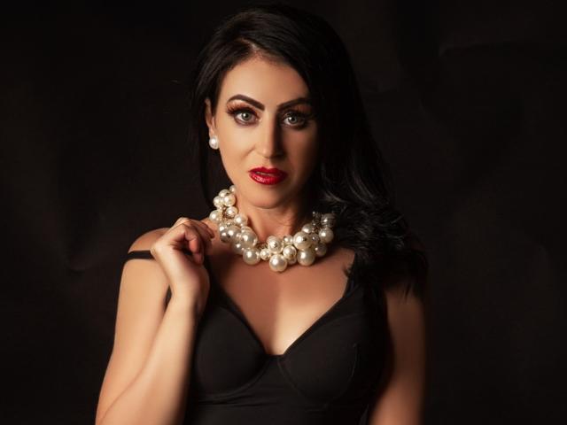 MistressTaylor with black hair, red lipstick, pearl necklace, and black dress, holding strap, showing her tits, dark background, sexy, confident.