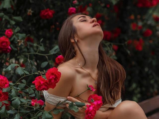 Karamel69 com cabelo castanho, olhos fechados, cabeça inclinada para trás, blusa branca com um ombro à mostra, segurando rosas vermelhas, expondo seus pequenos seios, cercada por folhas verdes e pétalas de rosas vermelhas..