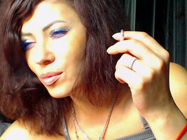 JuliaIce, con i capelli castano ondulati, il trucco occhi blu e un anello alla mano destra, che mostra la lingua, indossa una maglietta grigia e una collana d'argento..