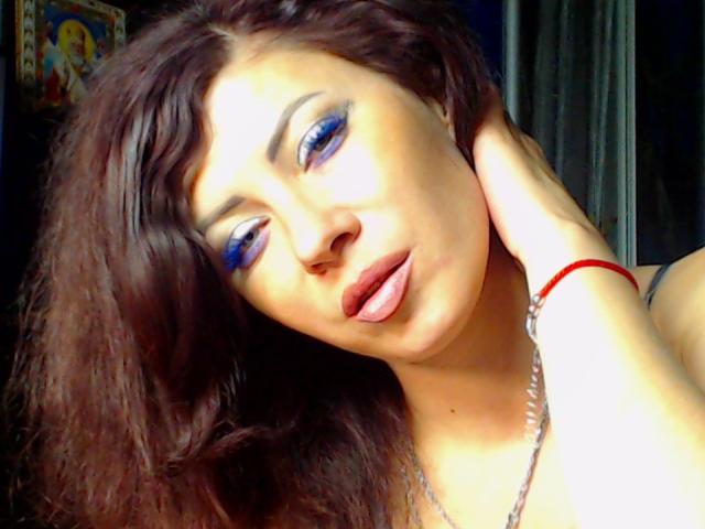 JuliaIce con sombra de ojos azul, cabello ondulado oscuro y lápiz labial rojo, con la lengua ligeramente afuera, una mano en el cabello, vistiendo una pulsera roja y un collar de plata..