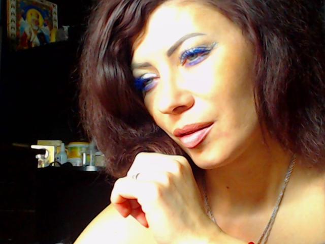 JuliaIce, con i capelli castano ondulati, trucco azzurro sugli occhi, labbra rosa e una collana d'argento, tocca il mento con un dito ornato da un anello. Il suo seno non è visibile..