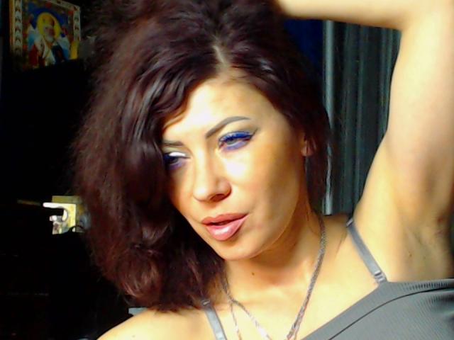 JuliaIce ha i capelli castano ondulati, il trucco occhi blu e un'espressione accigliata, con un braccio sollevato e una canotta grigia che mostra il seno e una collana d'argento..