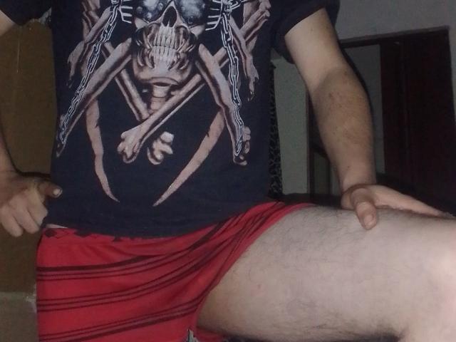 BigWalter con una camiseta negra con estampado de calavera, pantalones cortos rojos, mostrando un muslo cubierto de vello con una mano sobre la pierna, ligeramente flexionada, en una habitación con poca luz. Las manos descansan sobre las rodillas, visibles desde la cintura hacia abajo..