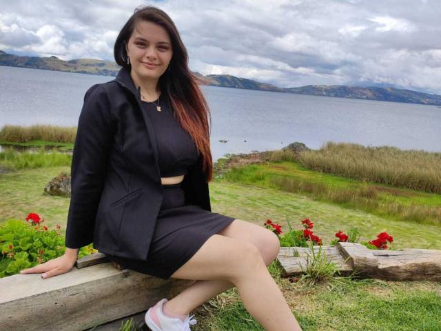 Katherin, con lunghi capelli castani, un blazer nero e una gonna nera, seduta su una recinzione di legno, che mostra le cosce, indossa scarpe da ginnastica bianche, sorride, vicino a un lago..