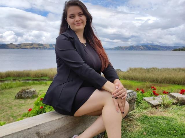 Katherin, una ragazza con lunghi capelli castani, vestita con un abito nero e una giacca, seduta su una panca di legno, sorridente, con le gambe incrociate e le mani sulle ginocchia, che mostra il sedere e le unghie dipinte di nero..