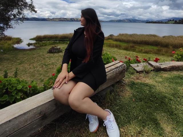KatherinLatin with long red-tinted hair, black dress, and white sneakers, sitting on a wooden bench, legs crossed, hands on knees, looking to the side, by a grassy lake.