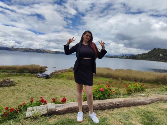katherinLatin with tits in a black top and skirt, hands up, standing on grass, log in front, red flowers, white sneakers, cloudy sky, lake in background.