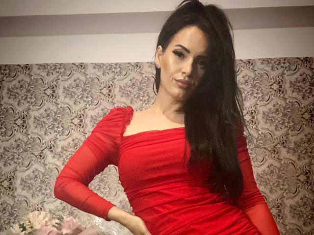 MissGabrielleVonD mit langen schwarzen Haaren, rotem Kleid und einer Hand am Hüfte, steht selbstbewusst da, zeigt ihr Gesäß und ihre kleinen Brüste und lächelt leicht..