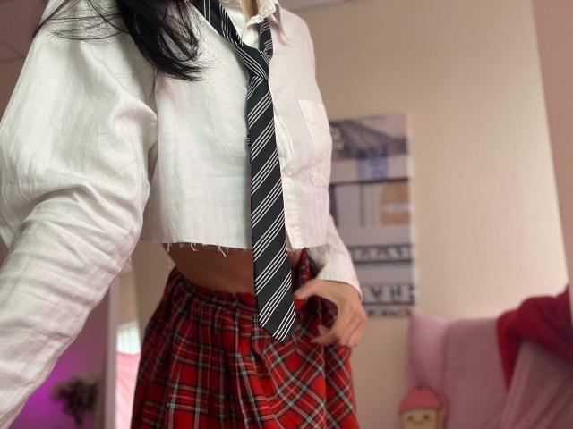 Tsukerberg with black hair, white crop top, and striped tie, wearing red plaid skirt, showing a flat stomach, hands on waist, in a softly lit room.