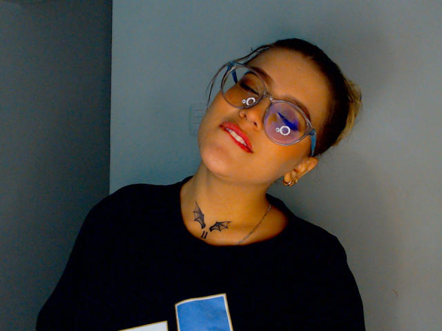 Kim, en été, avec des lunettes, du rouge à lèvres rouge, une chemise noire, un tatouage de chauve-souris sur le cou, tenant un téléphone, la tête penchée, et une expression légèrement amusée..