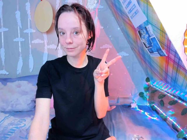 MissAstravert with wet brown hair, wearing a black shirt, sitting on a bed, flashing a peace sign with her right hand, looking cute and relaxed.
