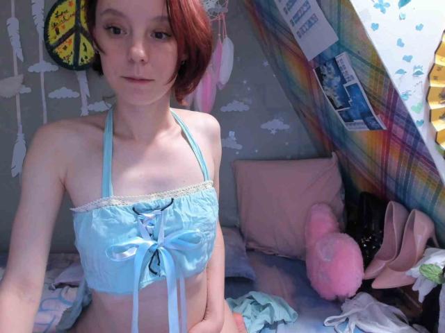 MissAstravert, avec ses courts cheveux roux, portant un haut bleu à lacets qui découvre sa petite poitrine et sa peau pâle, se tient dans une chambre en désordre avec des coussins roses..