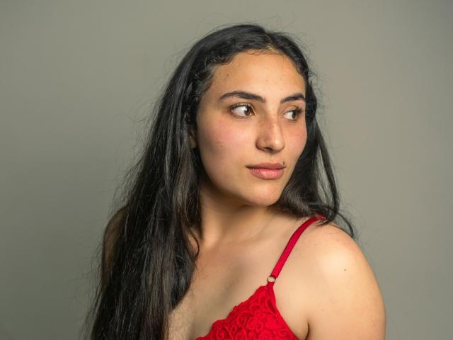 HelaViKy, con cabello largo y negro, un sujetador de encaje rojo, mirando hacia un lado, con una ligera sonrisa, ojos marrones, piel clara y un ligero rubor. Sus pechos son pequeños y visibles..