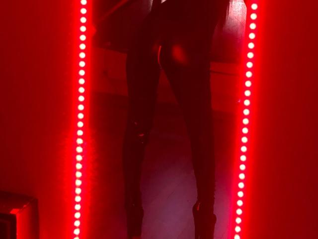 Il sedere e le cosce scoperte di MistressDonnaX69 sono illuminate da luci LED rosse, mentre è in piedi in una stanza buia con le gambe leggermente divaricate e indossa tacchi alti neri..