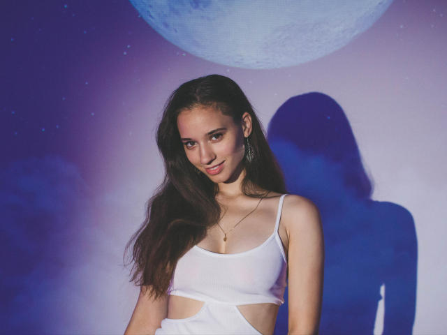 StacieLove with long dark hair, white crop top, small tits, and a gold necklace, smiling at the camera, shadowed against a purple and blue background.