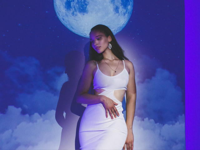 StacieLove with dark hair, white crop top, and white skirt, standing, hand on her ass, moon and clouds behind, purple and blue lighting, wearing large earrings, looking to side.